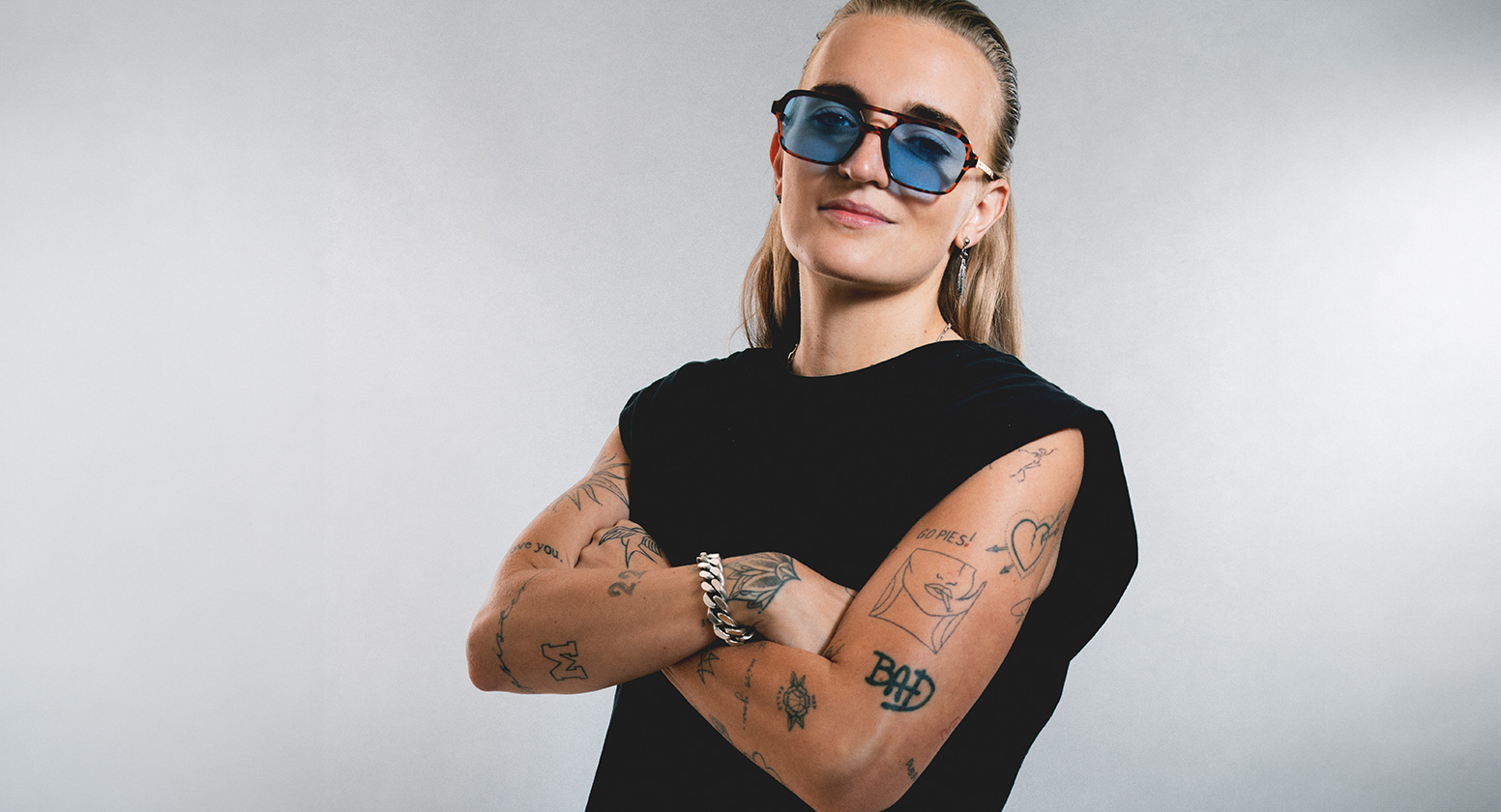 G Flip stands with a confident stance, wearing a black t-shirt and blue jeans, stands against a grey background. They sport various tattoos on their arms, sunglasses, and a silver chain bracelet.