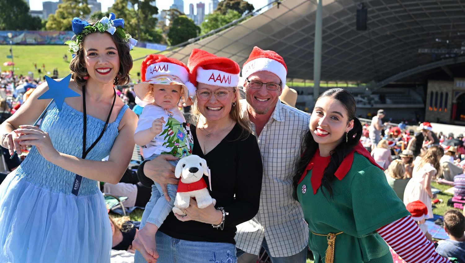 A man, a woman and a child wear Santa hats between two women dressed as a fairy and an elf in front of the Sidney Myer Music Bowl during the day at Carols