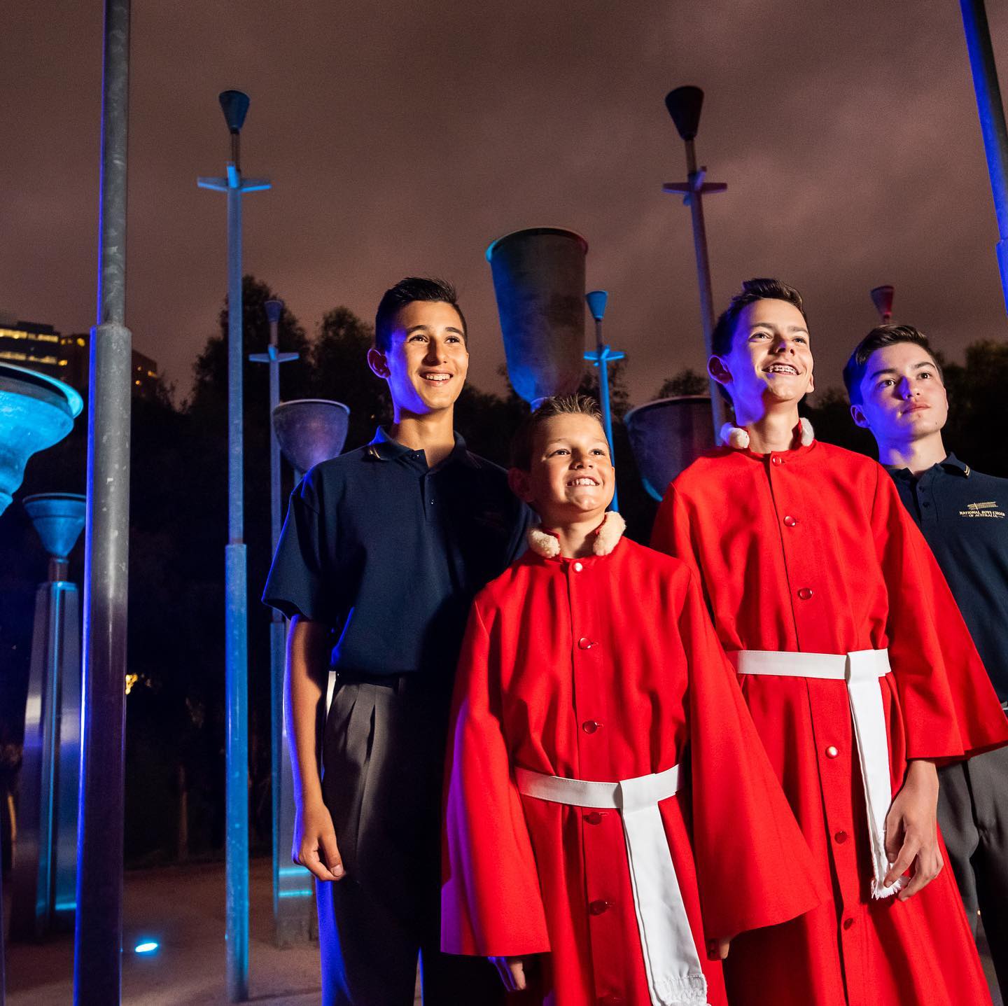 Four National Boys Choir members; two are wearing navy polo shirts and grey pants, and two are wearing red robes