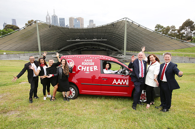Members from the Melbourne Gospel Choir and Vision Australia staff excitedly stand on the driver's side of a red AAMI cheer van on the lawn of the Sidney Myer Music Bowl