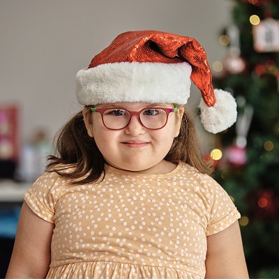 Daisy, 3, wears a santa hat and is standing in front of a Christmas tree