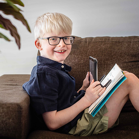Sam, aged 8, sits on a couch, holding a magnifier above a book and is looking at the camera