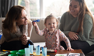 girl with low vision holding a fork, with mother and friend smiling at her
