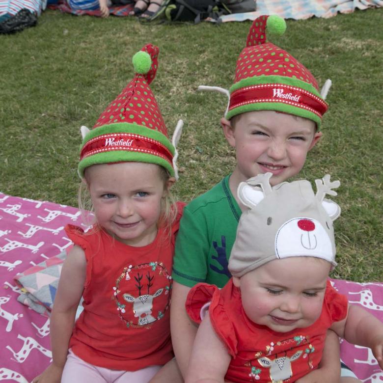 photo of three young children Carols guests smiling and wearing Christmas T-shirts and decorative Christmas hats.