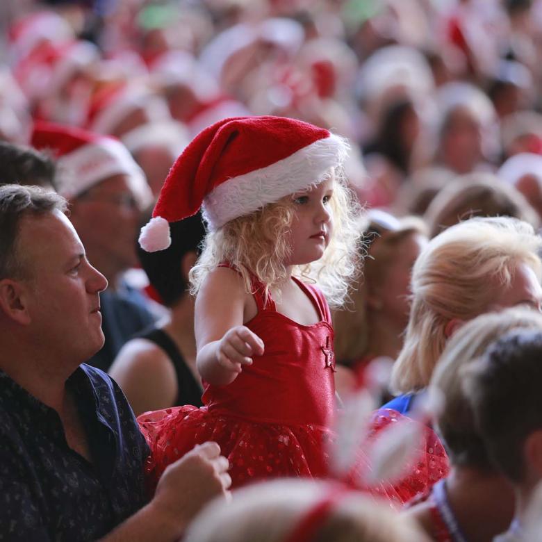 photo of the Carols crowd focusing on a father and daughter watching performances. The daugter is wearing a red outfit and a santa hat