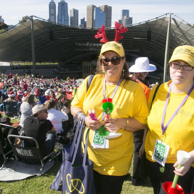 photo of two Carols volunteer event staff wearing yellow t-shirts standing with the crowd and stage behind them
