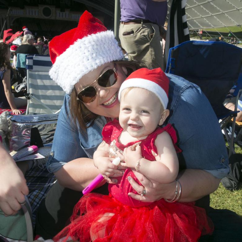photo of a woman wearing a santa hat and a baby wearing a red dress and a santa hat in her arms. Both look happy and are sitting on the grass