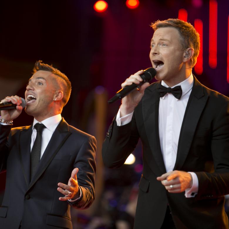 Photo of Anthony Callea and Tim Campbell singing together. Anthony Callea is wearing a black tie and Tim Campbell is wearing a black bowtie and both are wearing suits and singing into handheld microphones
