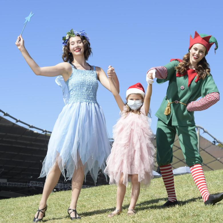 A fairy and an elf hold up the hands of a young girl in a santa hat