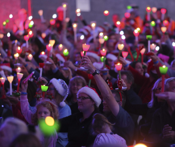  A large group of people sitting in the seated stalls section at Carols by Candlelight.  They are holding green and red candles and pink glowsticks up in the air, while they look towards the stage. A number of people are wearing Santa hats and Christmas accessories.