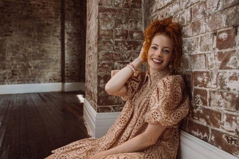 Emma Watkins leans against a brick wall smiling at camera