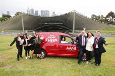 Members of the Melbourne Gospel Choir and Vision Australia staff excitedly stand on the driver's side of a red AAMI cheer van on the lawn of the Sidney Myer Music Bowl