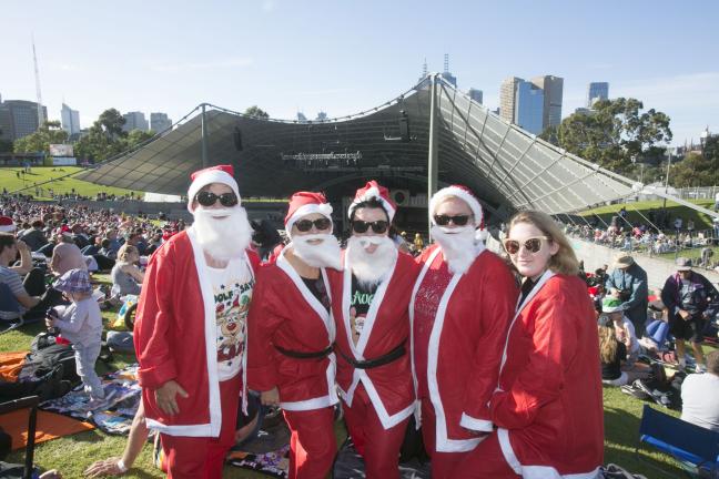 Five adults stand together on the lawn dressed in Santa costumes. The Sidney Myer Music bowl and crowd is behind them.