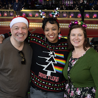 Three people wearing Christmas headwear stand in front of the Carols stage smiling with arms around eachother