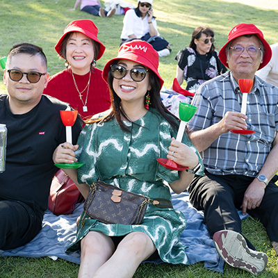 Five people sitting on a blanket on a lawn wear red bucket hats and hold candles smiling at the camera
