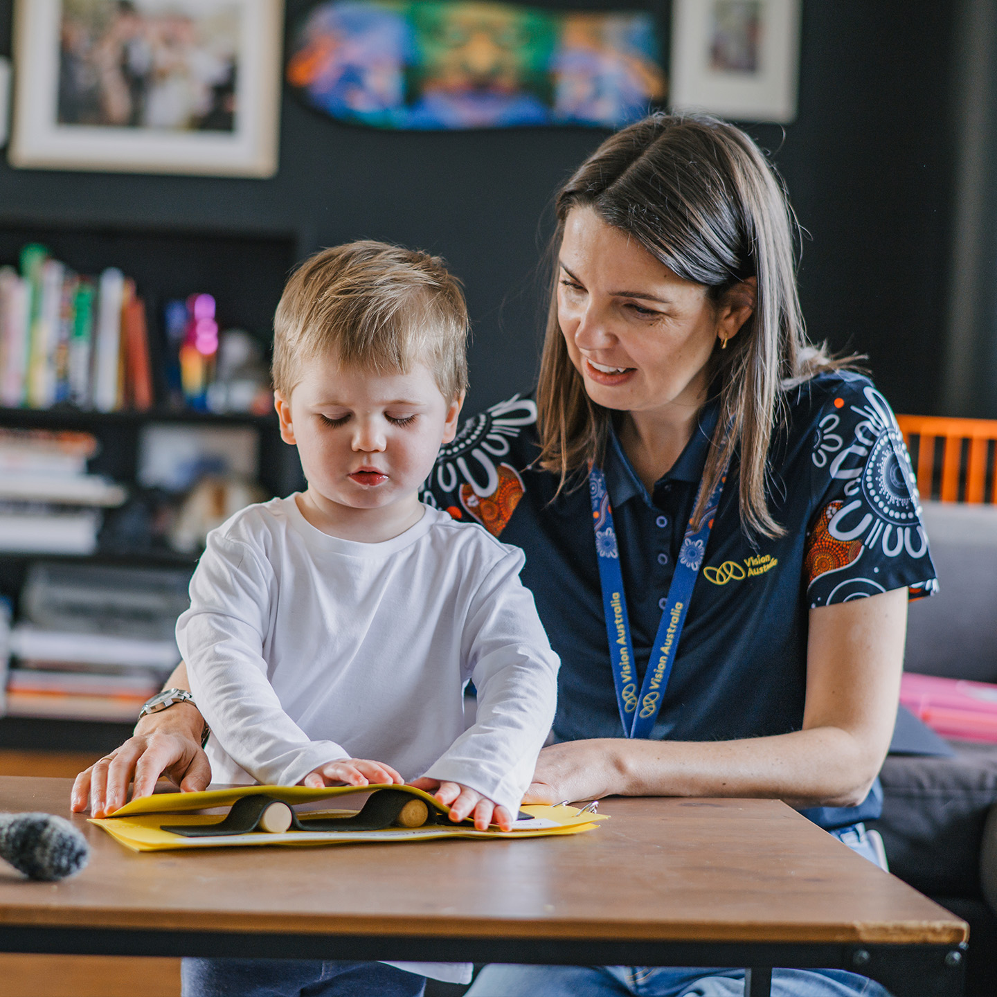 Young boy feels a book on the table sitting next to a woman in a Vision Australia uniform