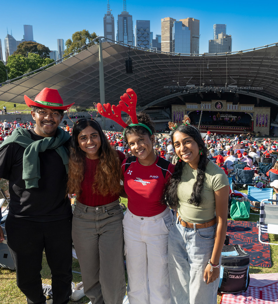 Three women and a man wearing Christmas headwear stand in the Carols crowd with the bowl in the background