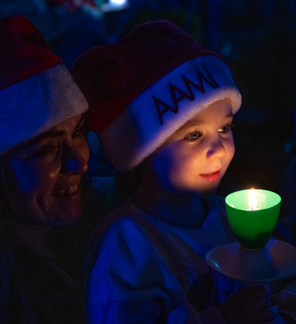 Child wearing Santa hat holds a candle with the candlelight on their face