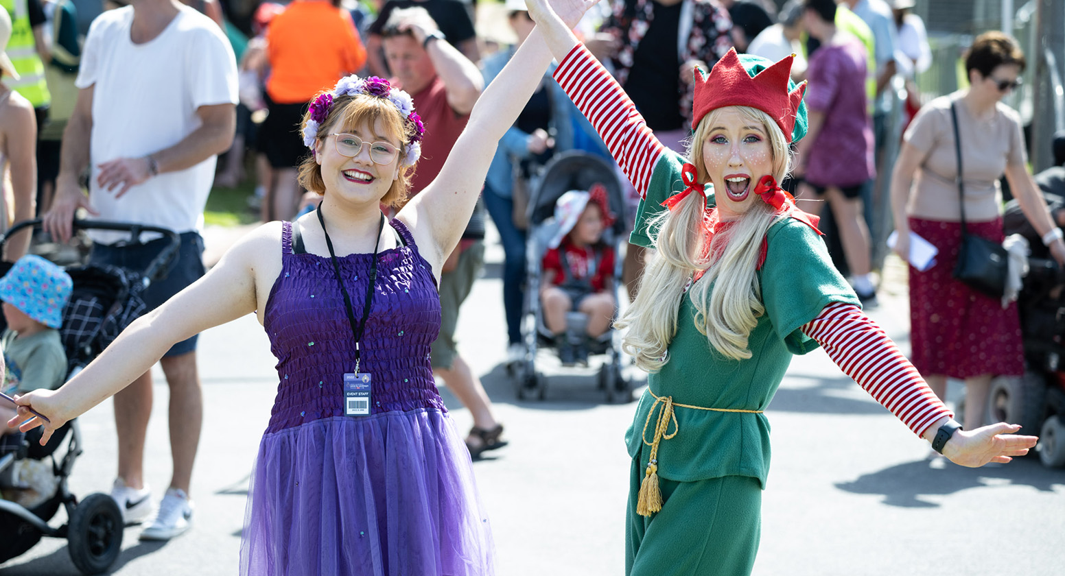 Two ladies, one dressed as an elf, have their hands up and smiling amongst a crowd