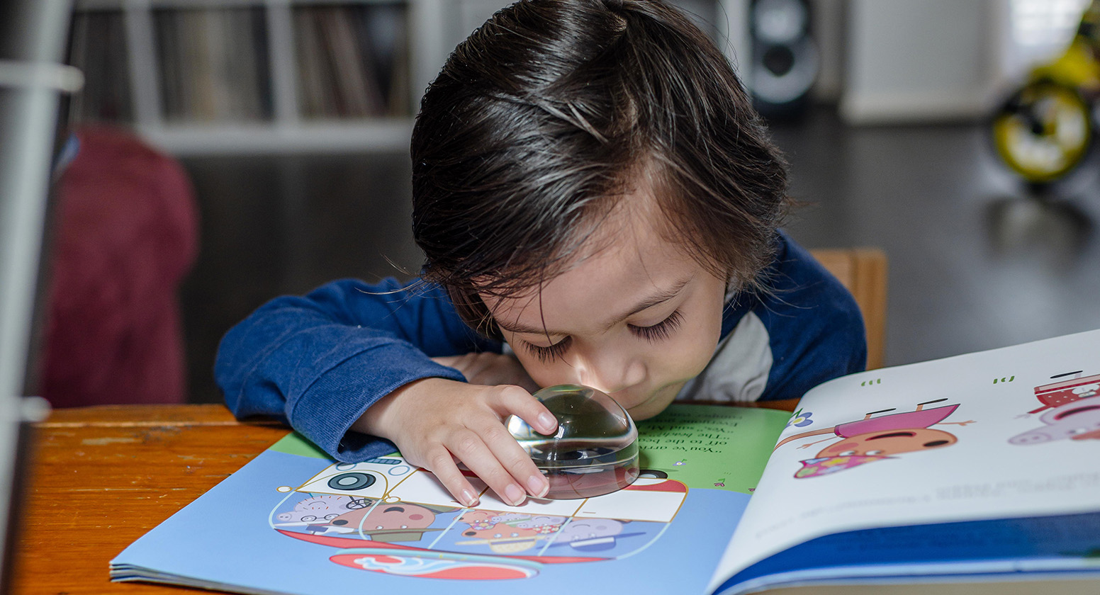 A young boy looks through a magnifier at a picture book on a table
