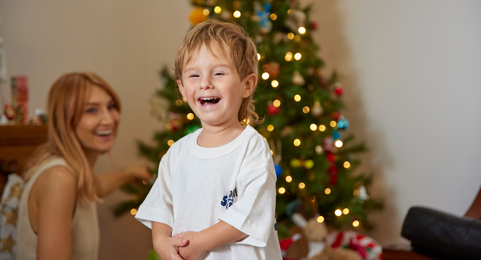 Young boy stands smiling in front of his mum sitting down next to a Christmas tree