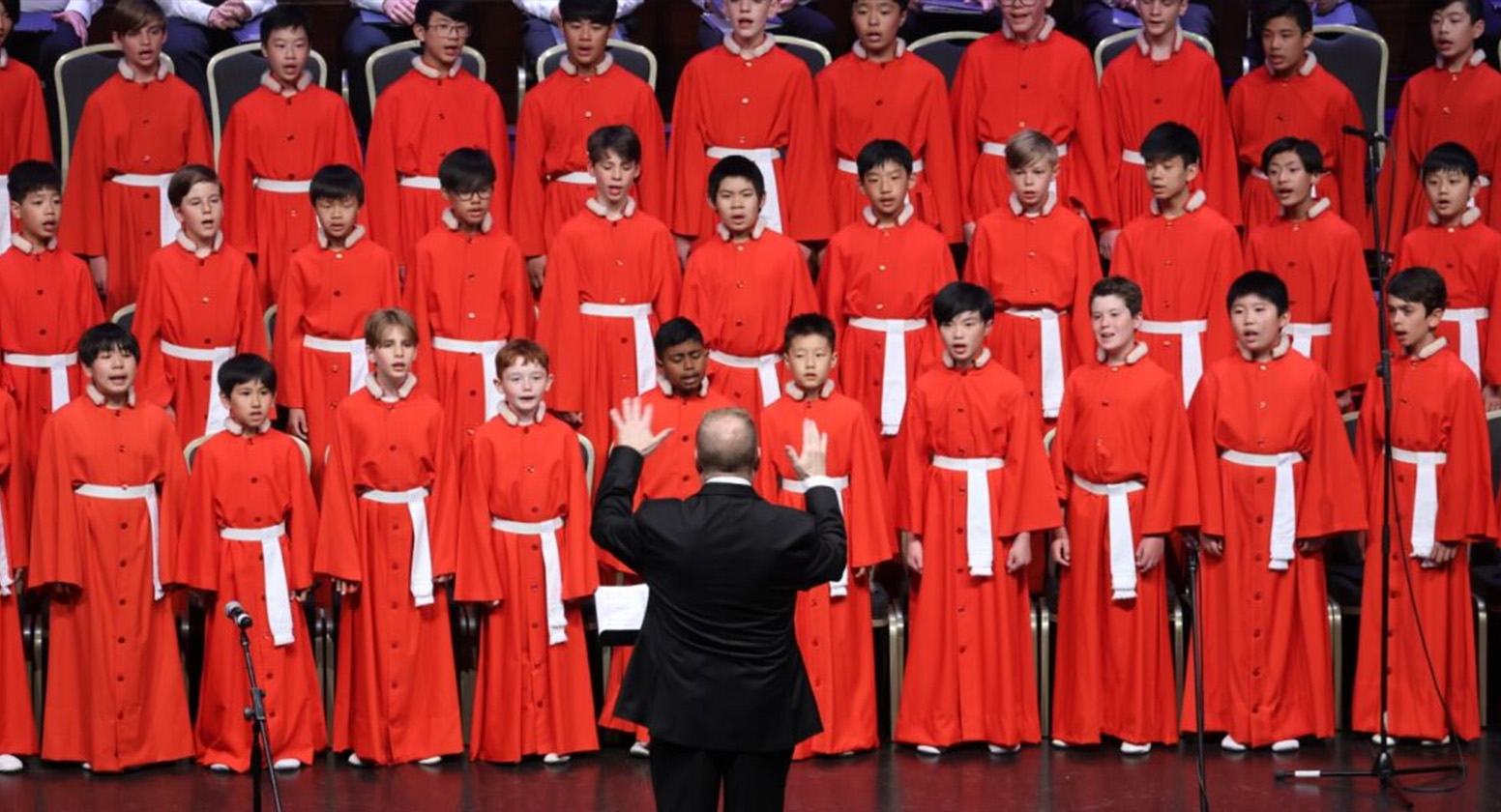 Boys in red robes with white sash belts stand in tiers singing, in front of a conductor
