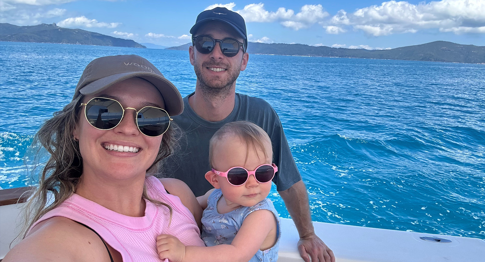 Woman holding young girl and man all wearing sunglasses take a selfie infront of water smiling