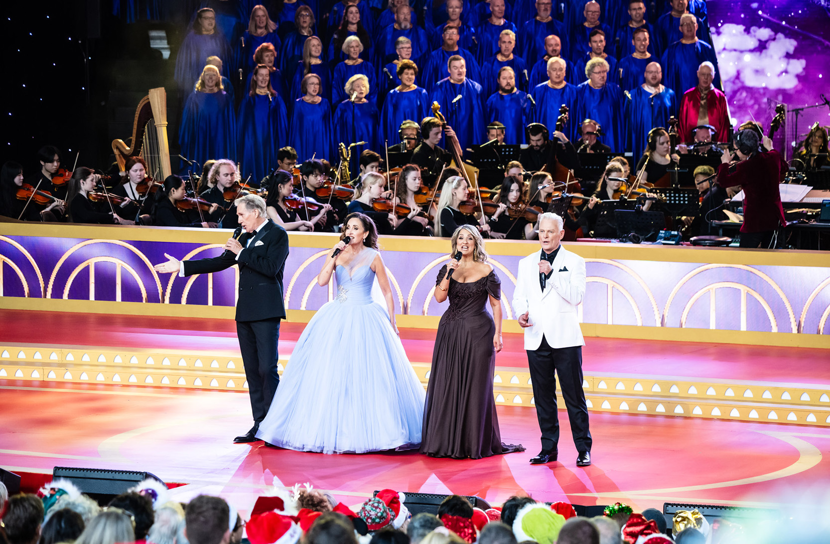 Four performers sing on the Carols stage with the choir behind them