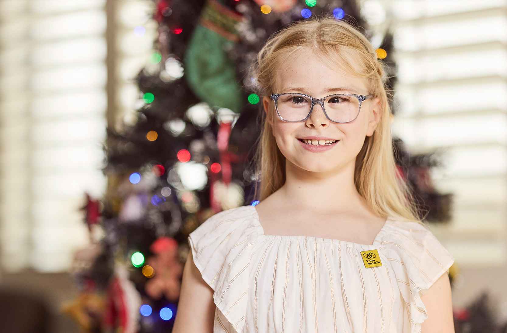 Young girl with blond hair, wearing glasses, stands in front of a Christmas tree