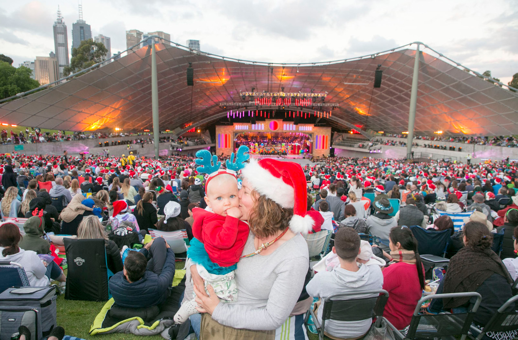 Woman wearing a santa hat kisses a baby with the carols crowd and stage behind them