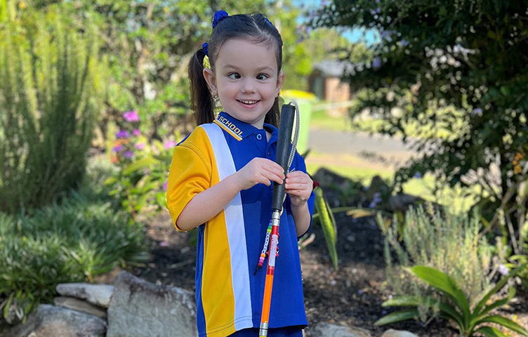 Young girl wearing yellow, blue and white polo stands outside smiling holding a cane