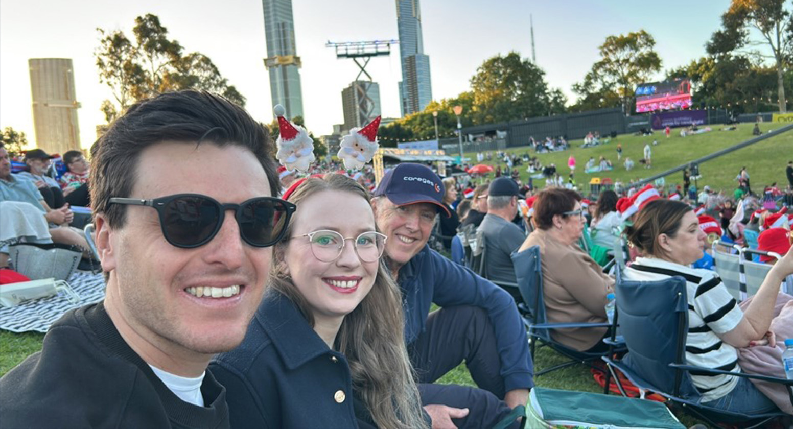 Two men and woman sit on the lawn at Carols by Candlelight smiling